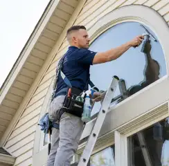 Professional window cleaner on a ladder using a squeegee to clean an exterior arched window, Aurora, IL.